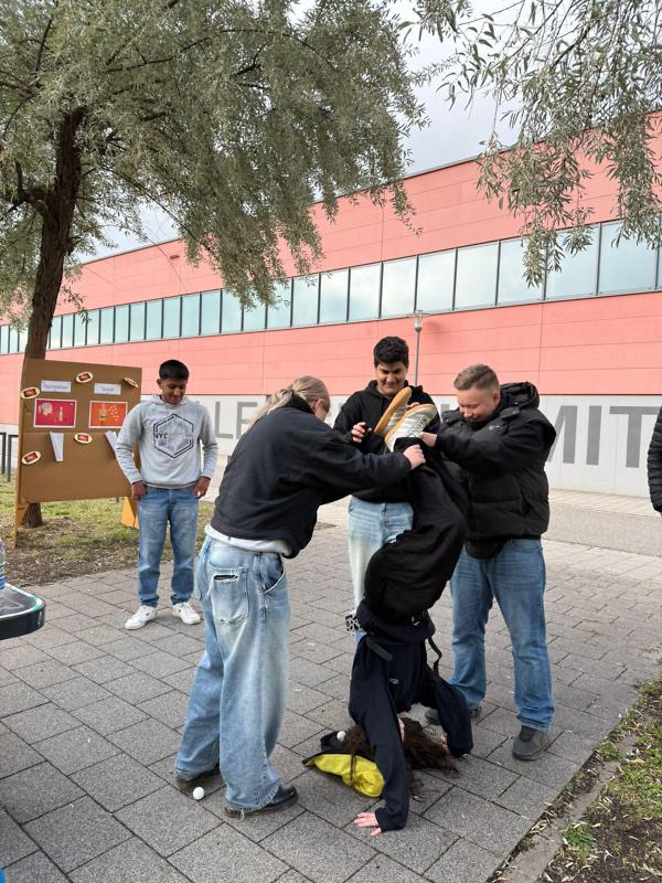 Fünf Auszubildende feiern gemeinsam ein Sommerfest. Einer von ihnen macht einen Handstand und wird von zwei anderen an den Beinen gesichert